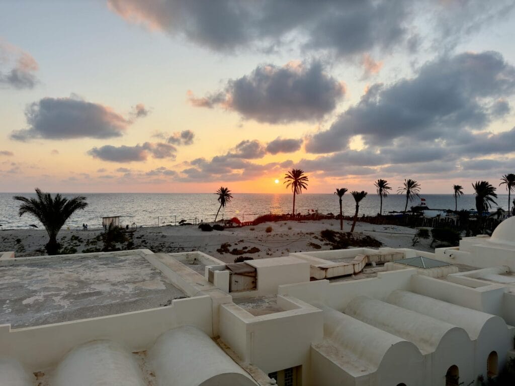 Un magnifique coucher de soleil sur une plage tunisienne avec des nuages épars dans le ciel. Le soleil, près de l'horizon, projette une lueur orange. Les palmiers se découpent sur le ciel et les élégants bâtiments blancs aux toits incurvés évoquent un endroit serein pour des vacances en villa.