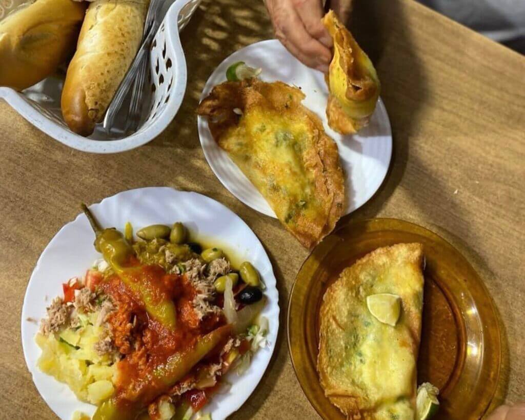 Une table avec une variété de nourriture : une assiette de chaussons farcis, un plat de légumes avec du chili et de la sauce, une corbeille de pain et une main tenant un chausson.
