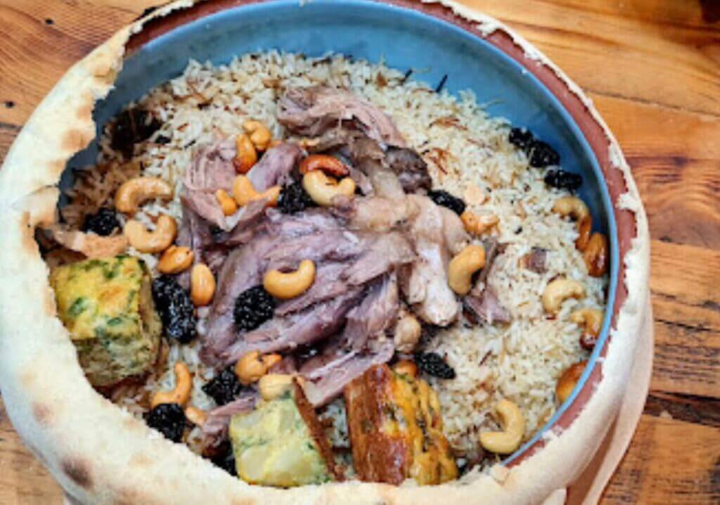 Un bol en céramique rempli de riz garni d'agneau râpé, de noix de cajou et de raisins secs est partiellement enfermé dans un pain de type pita. Des cubes de ce qui semble être un pain aux légumes ou aux herbes sont également visibles, le tout posé sur une table en bois.
