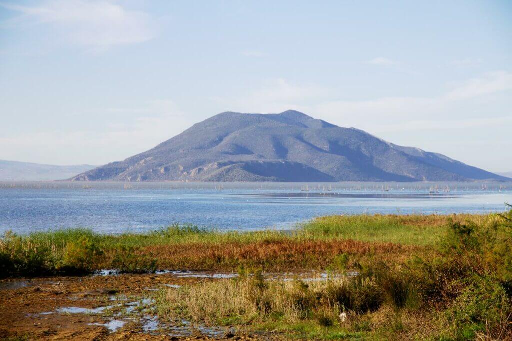 Un paysage serein au parc national d'Ichkeul en Tunisie présente une grande montagne au loin, bordée par un lac calme au premier plan. La scène comprend une végétation verte luxuriante et un ciel bleu avec des nuages légers, évoquant une atmosphère parfaite pour les amoureux de la nature en quête d'escapades paisibles.