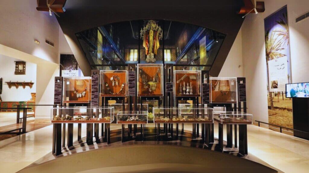 L'exposition du Musée du Patrimoine Traditionnel de Djerba présente des objets d'art dans des vitrines en verre, avec en arrière-plan un grand objet décoratif. Un éclairage complexe met en valeur des objets historiques, notamment des bijoux et des poteries, le tout dans un intérieur moderne et élégant.
