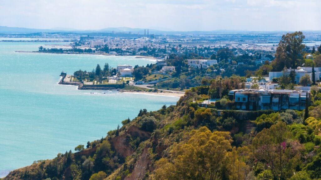 Une vue pittoresque sur la côte révèle une mer turquoise qui se courbe dans une baie bordée de plages de sable. Des collines verdoyantes, parsemées de villas et d'arbres, sont au premier plan. À l'arrière-plan se trouve un paysage urbain tentaculaire de bâtiments bas sous un ciel partiellement nuageux, parfait pour une aventure Dyarbnb.